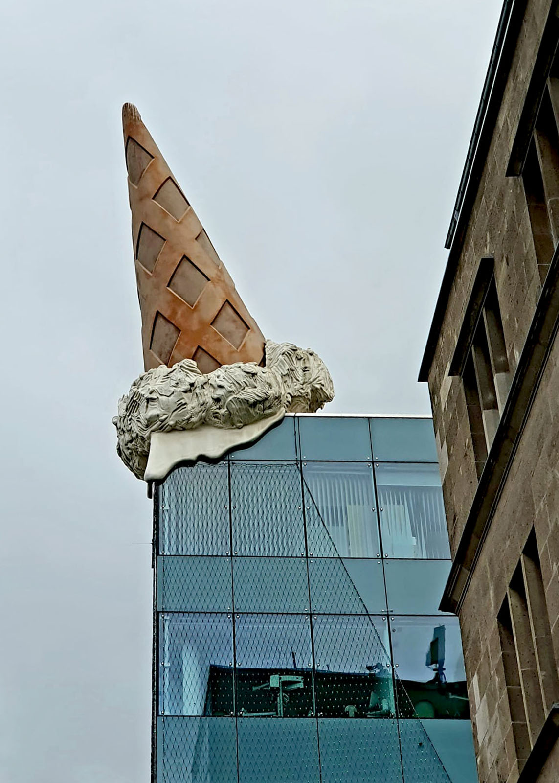Ein Gebäude mit Glasfront, über dessen Ecke eine übergroße Eiswaffel mit schmelzendem Eis gestülpt ist
