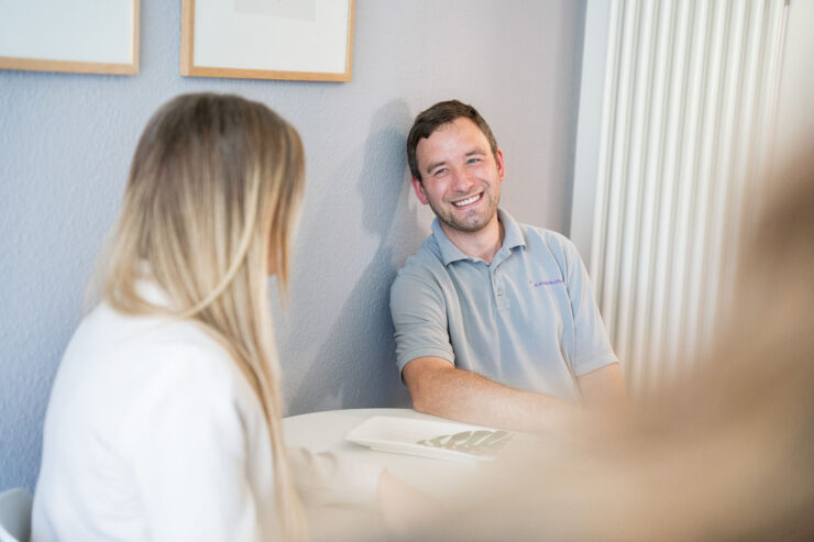 Ein junger Mann mit St. Elisabeth Stiftung Logo auf der Brust sitzt mit einer blonden Frau an einem Tisch und lächelt herzlich