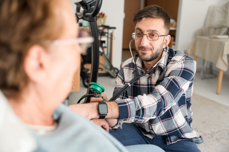 Ein Mann mit Brille misst den Blutdruck einer älteren Frau, die unscharf im Vordergrund zu erkennen ist