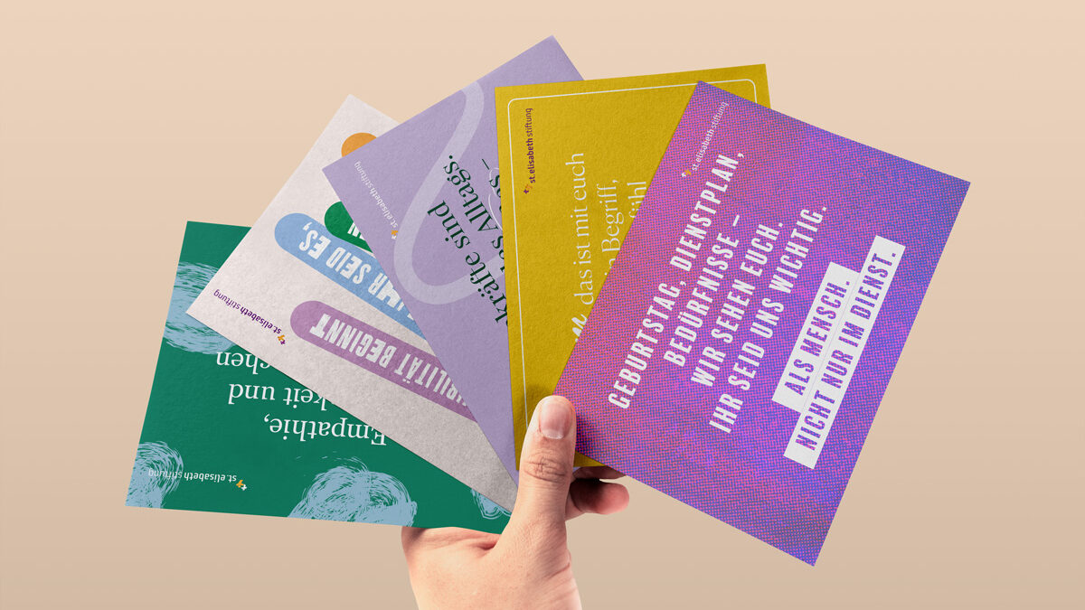 Eine Hand hält vier verschieden gestaltete Postkarten von der St. Elisabeth Stiftung in einem Fächer